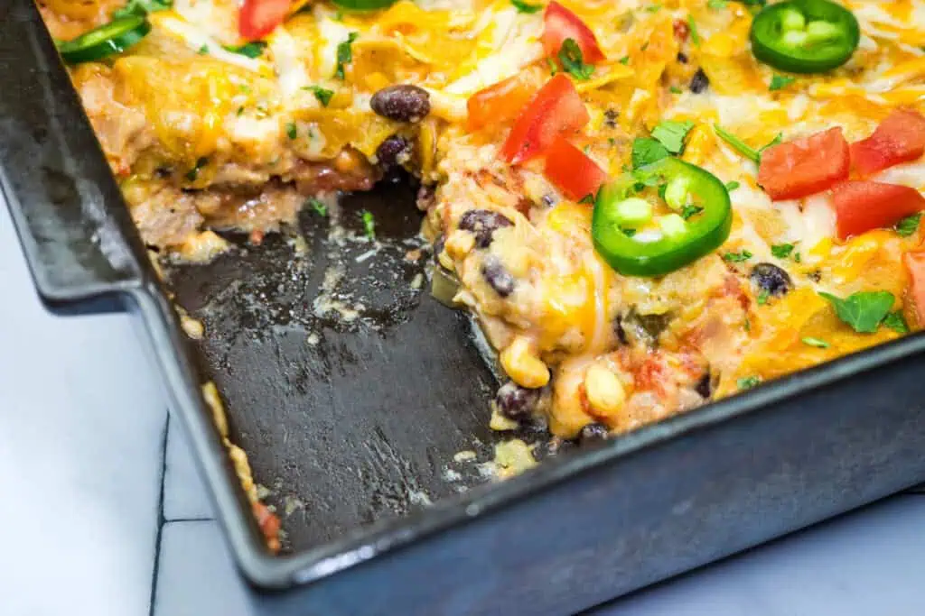 A Cheesy Mexican Casserole in a black dish, featuring layers of cheese, black beans, corn, and topped with jalapeño slices and diced tomatoes. A portion is missing from the corner.