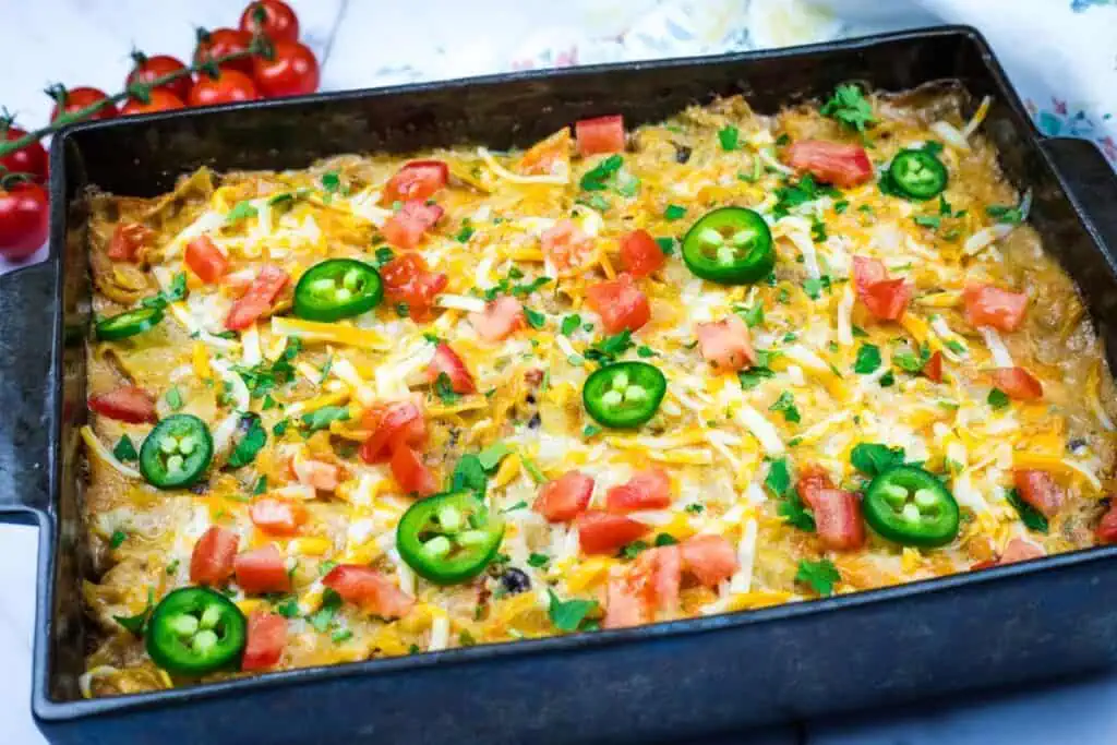 A Cheesy Mexican Casserole topped with sliced jalapeños, diced tomatoes, cheese, and herbs in a rectangular pan.