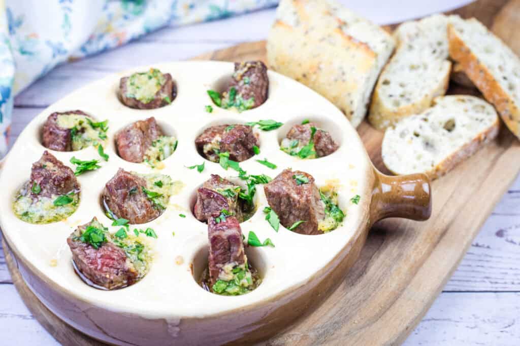 Ceramic dish with cooked escargot topped with parsley and butter, served alongside sliced bread on a wooden board.