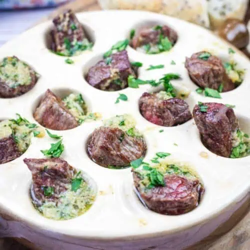 A bowl of garlic butter steak bites and bread on a wooden cutting board.