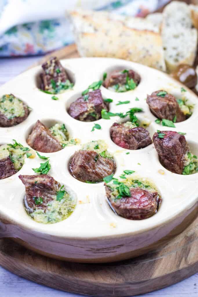 A bowl of garlic butter steak bites and bread on a wooden cutting board.