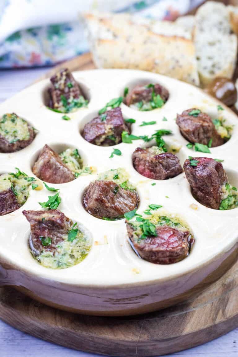 A bowl of garlic butter steak bites and bread on a wooden cutting board.