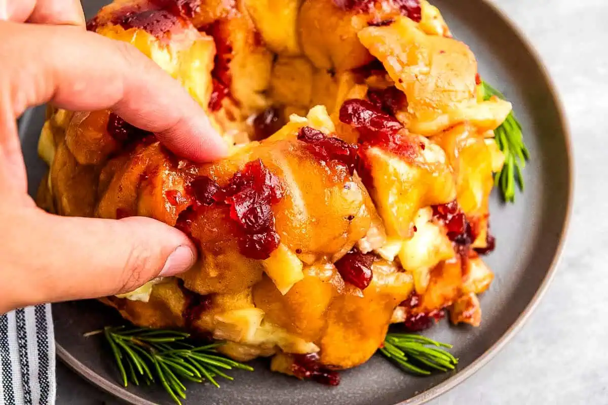 A hand pulls apart a Mini Cranberry-Brie Pull-Apart Bread garnished with rosemary, on a gray plate.