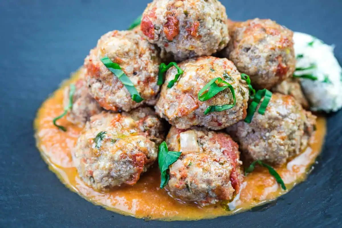 A stack of ricotta meatballs with tomato sauce, garnished with sliced green herbs, on a dark plate.