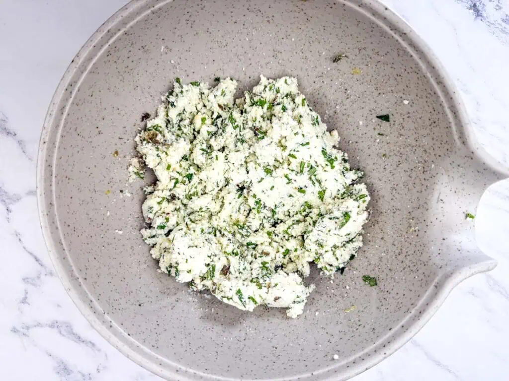 Combined ricotta, ¼ cup parmesan cheese, parsley, chives, basil, lemon zest, capers, garlic powder, onion powder, and red pepper flakes in a bowl on a white marble countertop.