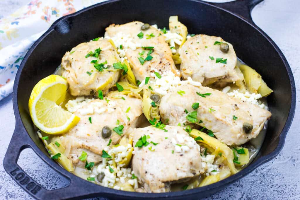 Chicken pieces with artichokes, capers, and herbs in a black pan, garnished with lemon wedges and chopped parsley.