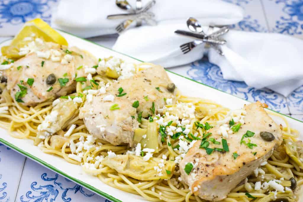 Plate of chicken piccata with pasta, garnished with capers, parsley, and cheese, served with lemon wedges. Napkins with decorative holders in the background.
