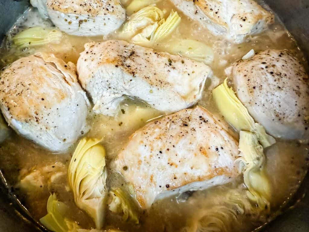 Several seasoned chicken breasts and artichoke pieces simmering in a pot of broth.