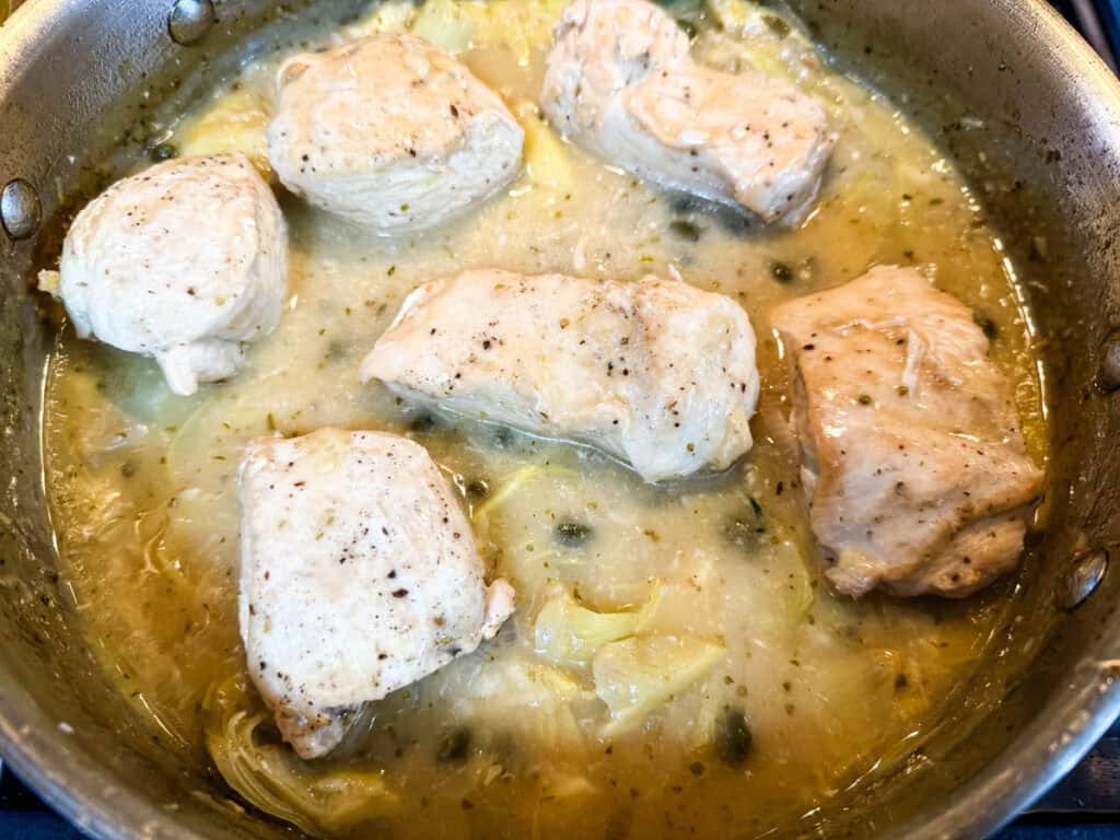Chicken pieces cooking in a pan with a light sauce and sliced onions.