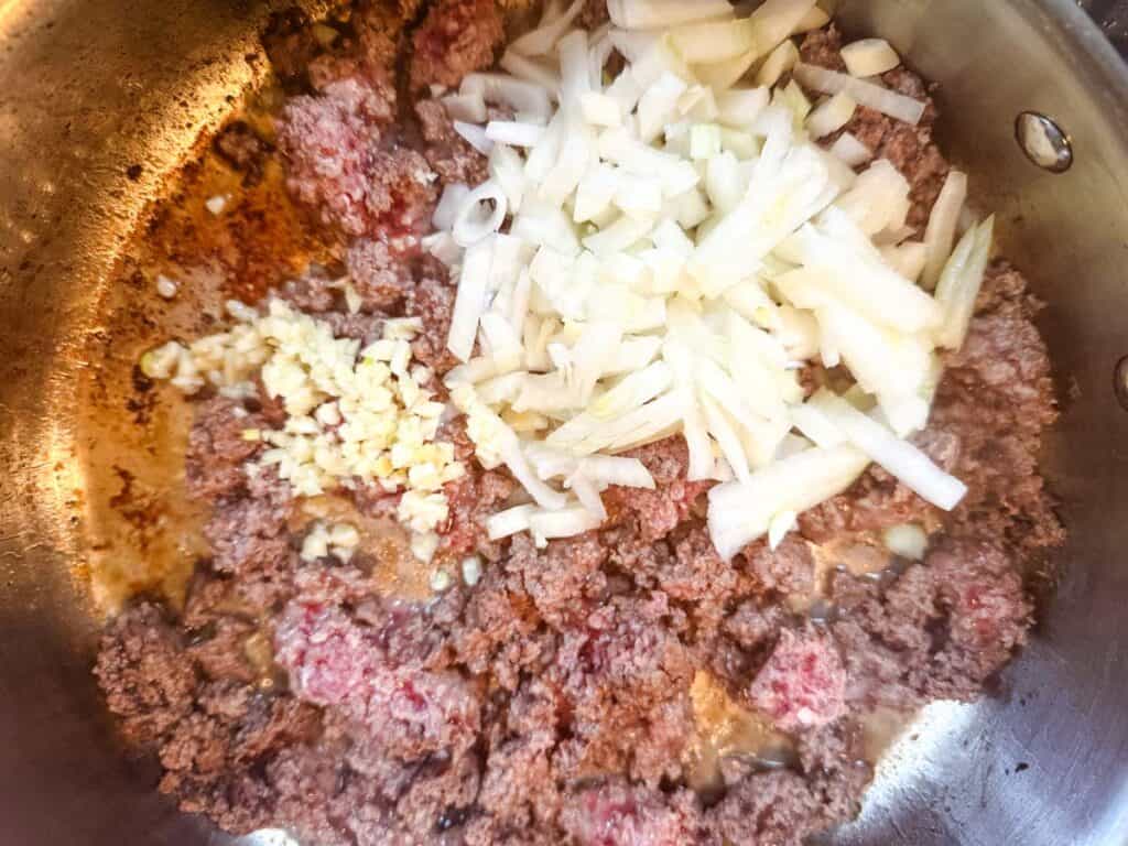 Crumbles of ground beef with diced white onions and minced garlic in a skillet.