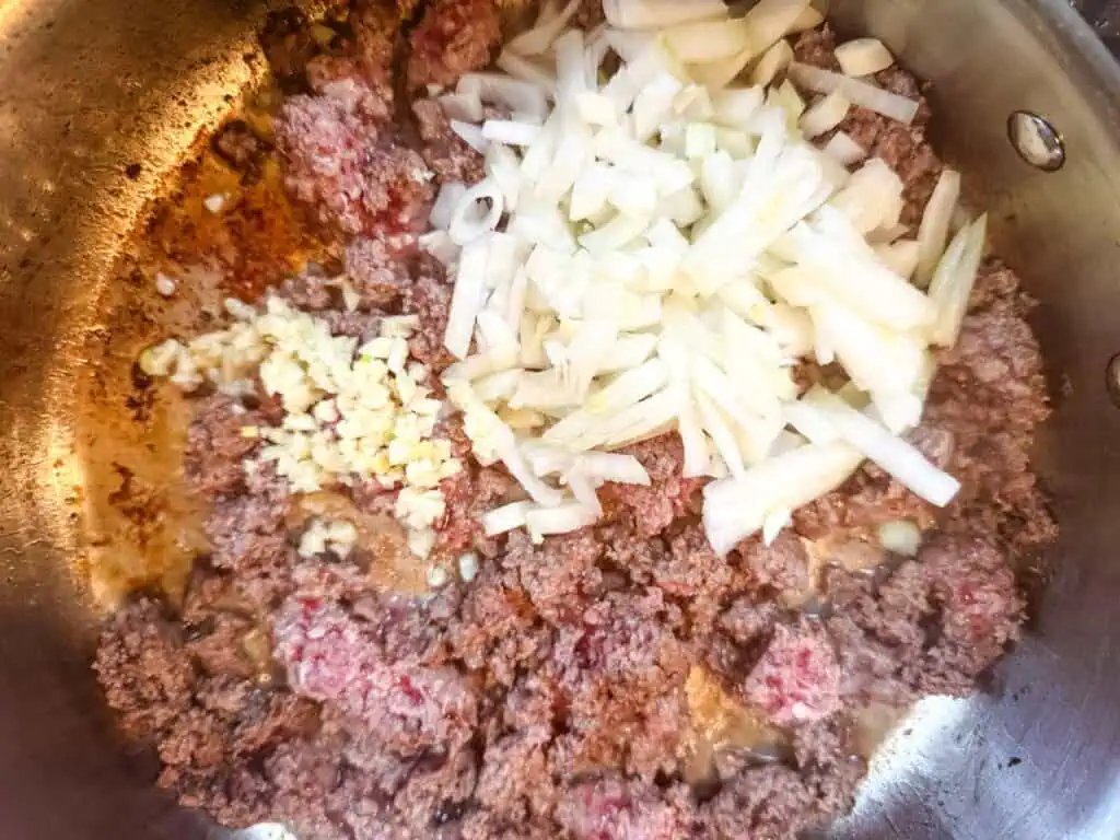 Crumbles of ground beef with diced white onions and minced garlic in a skillet.