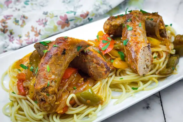 Sausages on spaghetti with bell peppers, garnished with herbs, served on a rectangular white plate.