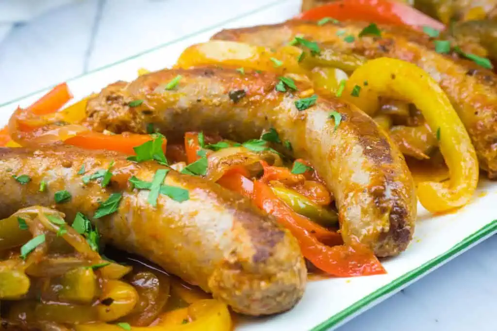 Close-up of cooked sausages mixed with sliced red and yellow bell peppers, garnished with chopped herbs on a white plate.