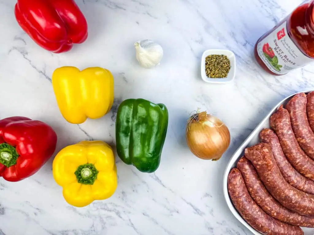 Assorted bell peppers, garlic, onion, oregano, a bottle of tomato sauce, and a tray of sausages are arranged on a marble surface.