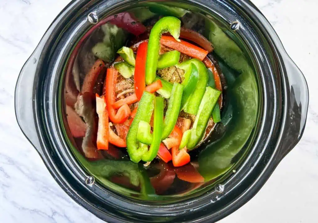 Sliced red and green bell peppers on top of sausages in a slow cooker on a white marble surface.