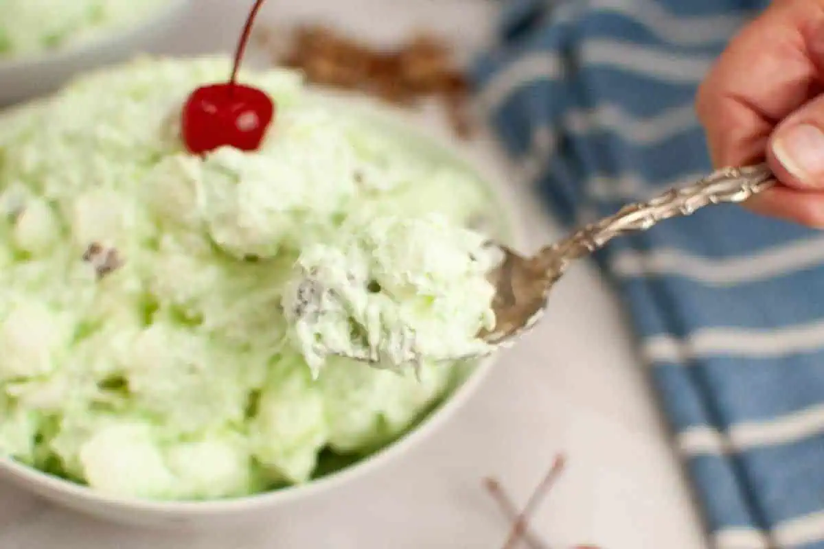 A spoonful of Watergate salad garnished with a cherry, is held over a bowl.
