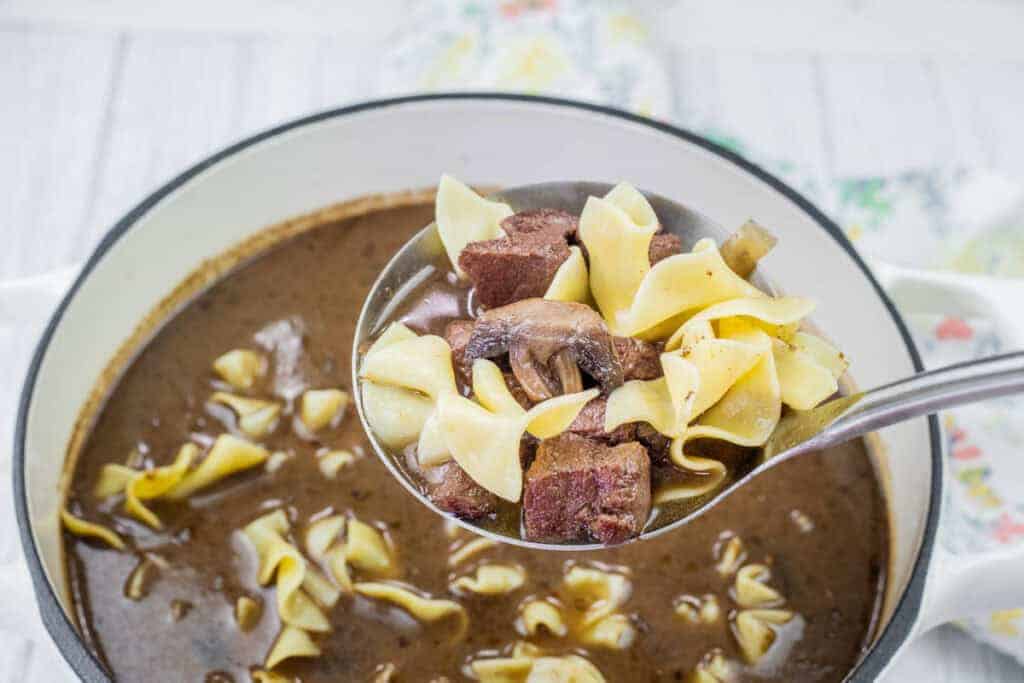 A ladle holds beef chunks, mushrooms, and egg noodles over a pot of Beef Stroganoff Soup.