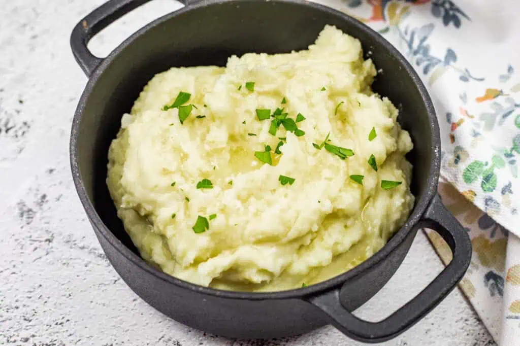 Rustic mashed potatoes served in a black pot, topped with melted butter and fresh parsley.