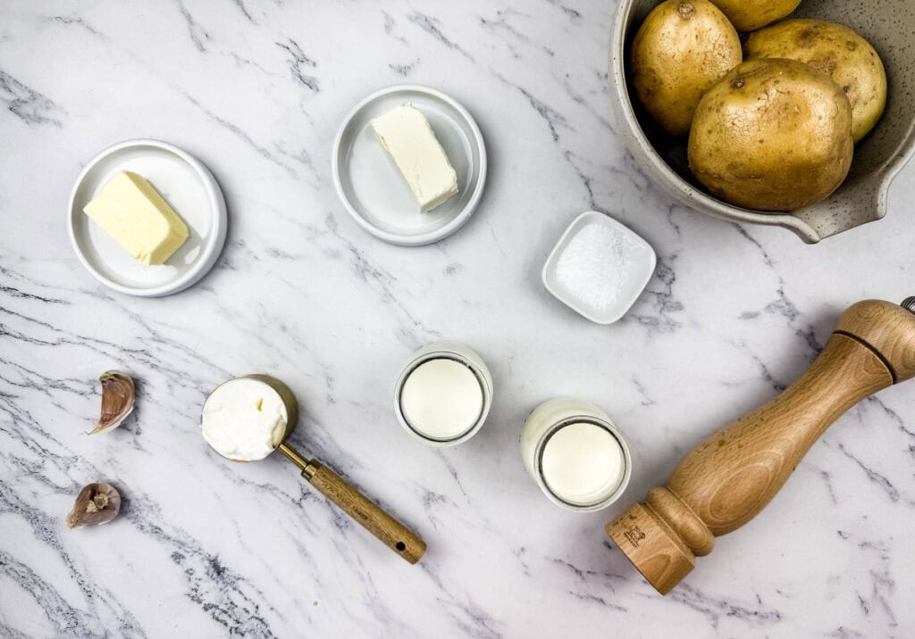 Mashed potato with cream cheese ingredients on a marble surface, including butter, cream cheese, sour cream, garlic, milk, salt, pepper, and a bowl of potatoes.