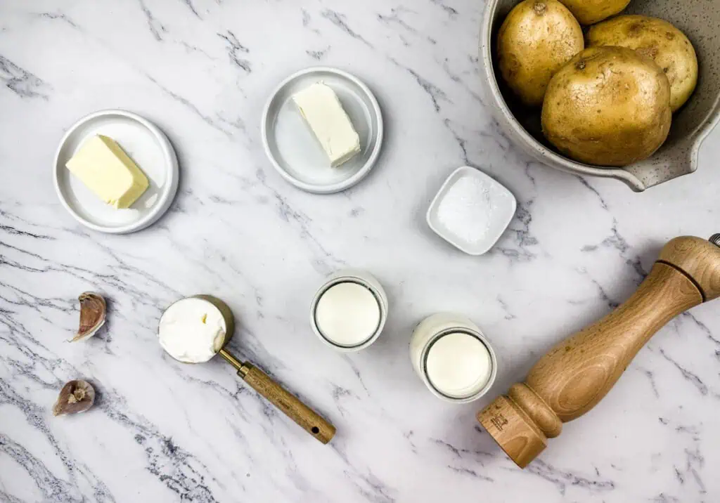 Mashed potato with cream cheese ingredients on a marble surface, including butter, cream cheese, sour cream, garlic, milk, salt, pepper, and a bowl of potatoes.