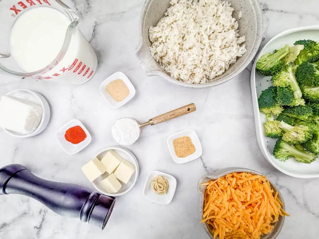 Ingredients for Broccoli Rice Casserole with Fresh Broccoli arranged on a marble countertop, including cooked rice, fresh broccoli, shredded cheddar cheese, butter, milk, cream cheese, flour, and seasonings.