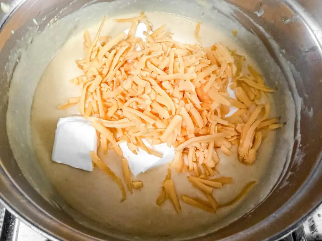 Shredded cheddar cheese and cream cheese added to a creamy sauce in a stainless steel pot, ready to be melted and combined.