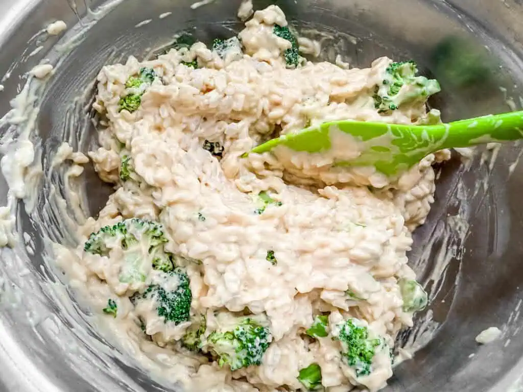 Broccoli, rice, and creamy cheese sauce being mixed together in a stainless steel bowl with a green silicone spatula, preparing for casserole baking.