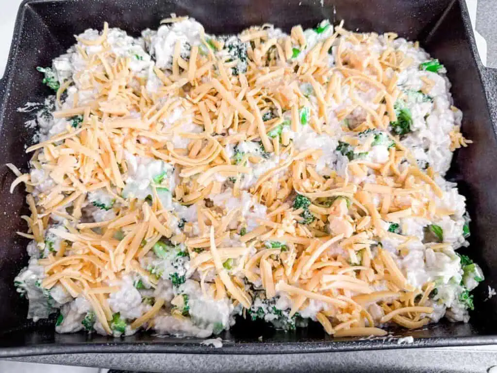 Unbaked broccoli rice casserole in a black baking dish, topped with shredded cheddar cheese, ready to be placed in the oven.