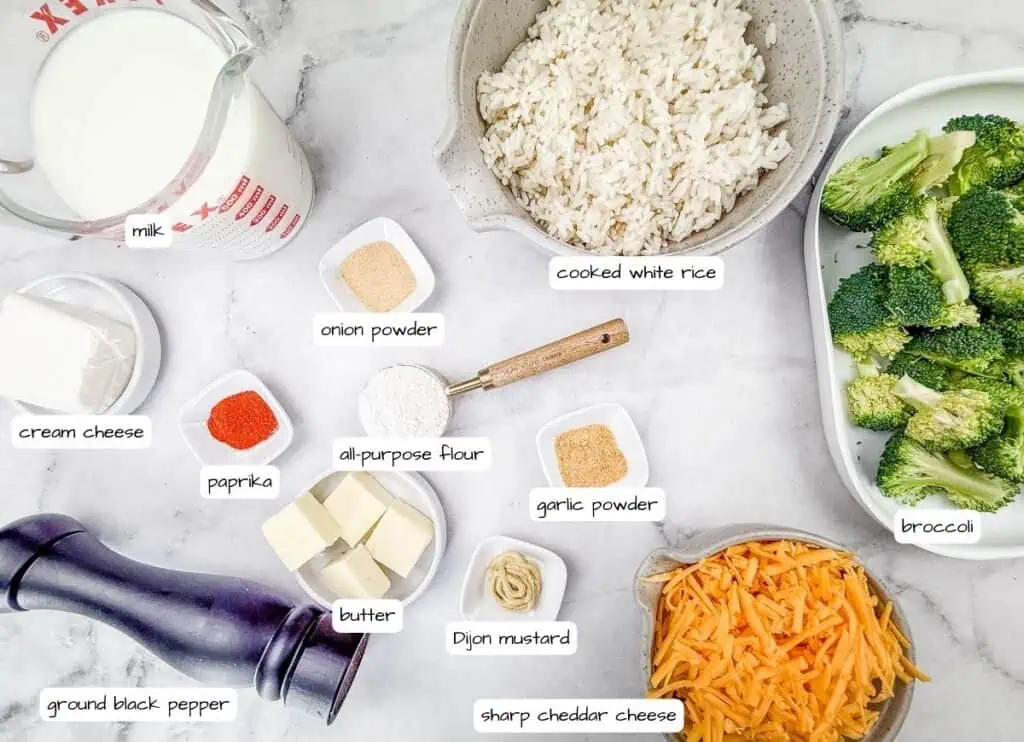 Ingredients for Broccoli Rice Casserole with Fresh Broccoli displayed on a marble countertop, including cooked white rice, fresh broccoli, sharp cheddar cheese, milk, cream cheese, butter, all-purpose flour, Dijon mustard, paprika, garlic powder, onion powder, and ground black pepper.