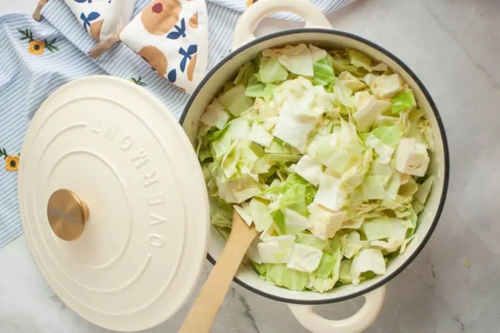 A white pot filled with chopped cabbage, a wooden spoon resting inside, with a striped cloth and oven mitt in the background.