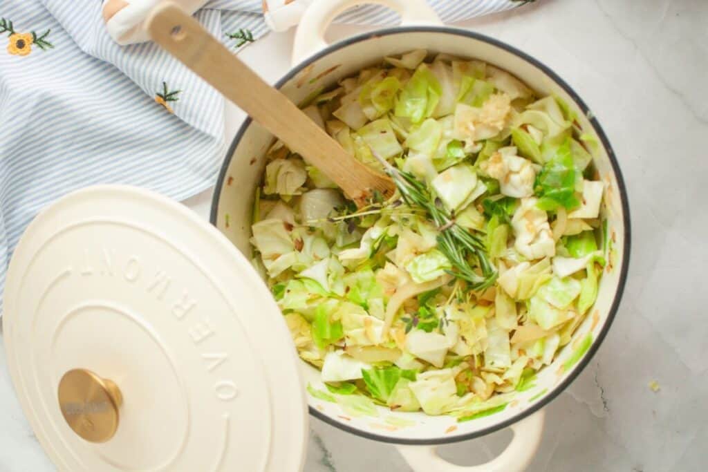White pot filled with chopped cabbage and herbs, placed on a striped cloth. A wooden spoon is inside the pot.