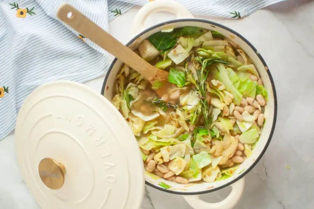 A pot of soup with cabbage, white beans, and herbs. The lid is partially open, and a wooden spoon rests inside the pot. The pot is on a striped cloth with daisy patterns.