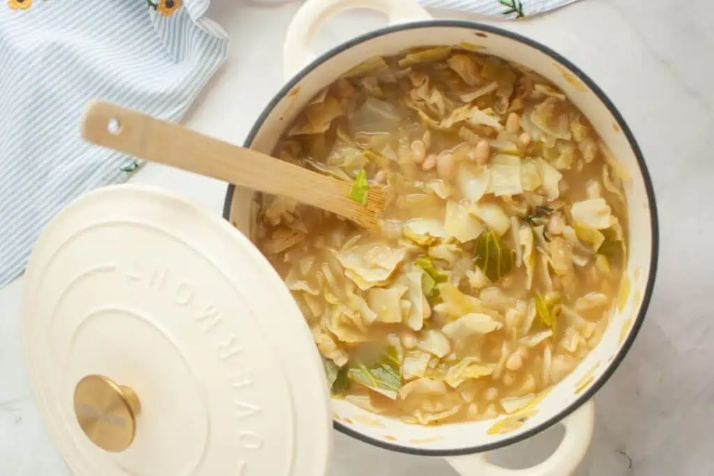 A pot of vegetable soup with cabbage, beans, and greens, accompanied by a wooden spoon. The pot lid rests partially open on a marble surface.