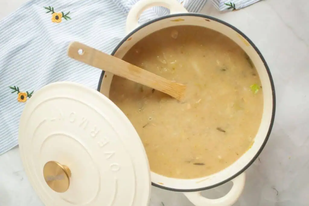 A pot of soup with a wooden spoon inside sits on a white surface next to a striped cloth with flower embroidery.