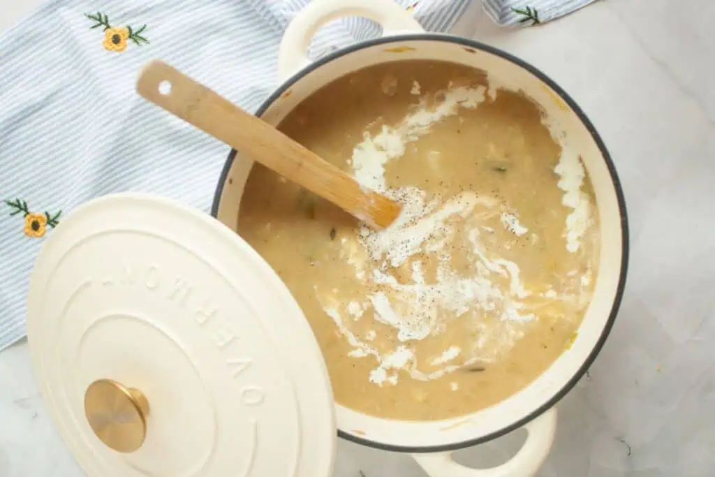 A pot of creamy soup with a wooden spoon, partially covered with a lid, on a striped cloth with embroidered flowers.