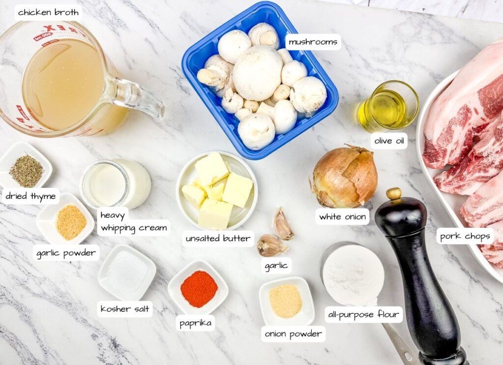 Ingredients labeled for Smothered Pork Chops with Onions & Mushrooms laid out on a marble surface, including chicken broth, mushrooms, pork chops, onion, spices, butter, cream, garlic, olive oil, and flour.