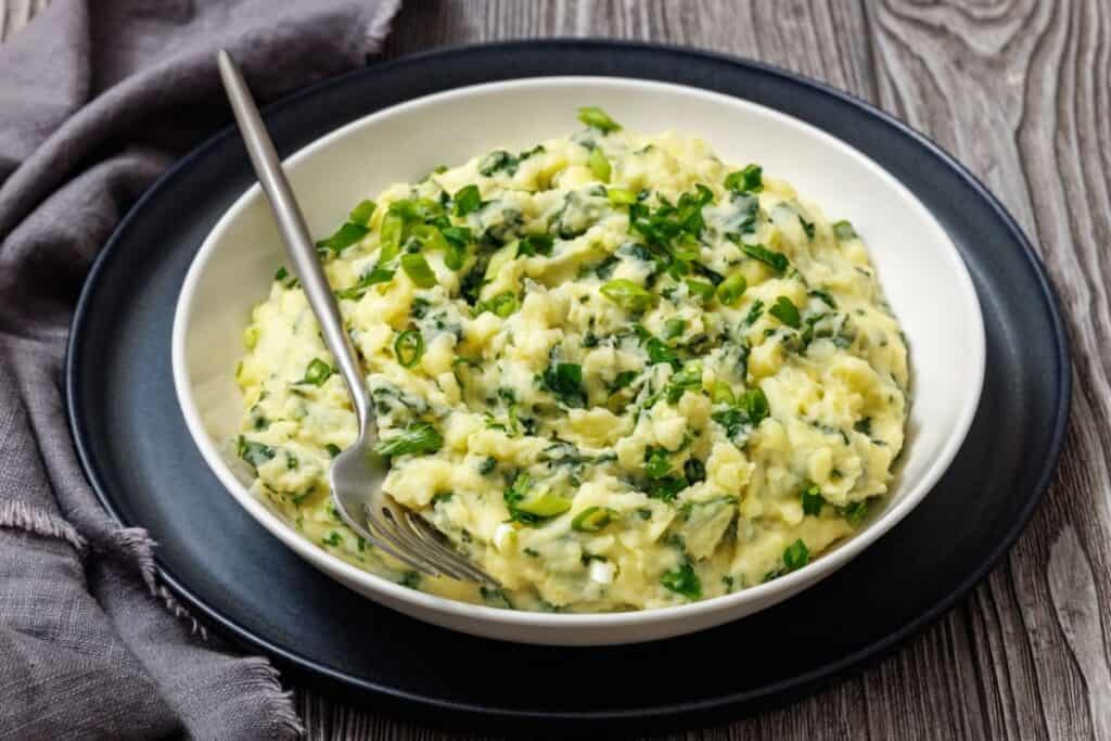A bowl of Colcannon with Kale, placed on a black plate with a fork in the dish, with a textured wooden background and grey cloth on the side.