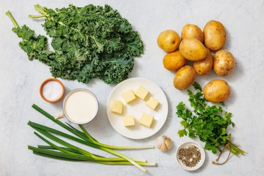 Ingredients for Colcannon with Kale on a surface: kale, potatoes, cubes of butter, garlic, green onions, parsley, a bowl of flour, a cup of milk, and a bowl of pepper.