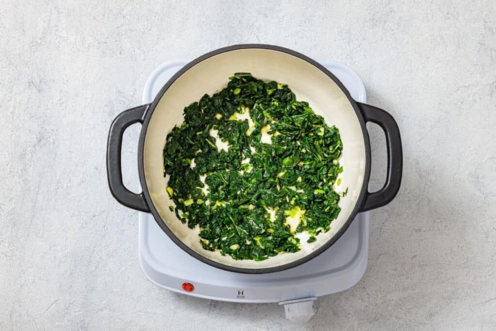 Chopped spinach being sautéed in a black pot on a white electric cooktop against a light gray background.