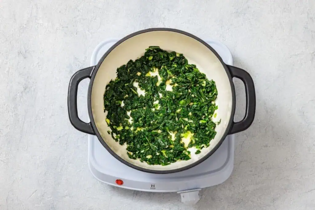 Chopped spinach being sautéed in a black pot on a white electric cooktop against a light gray background.