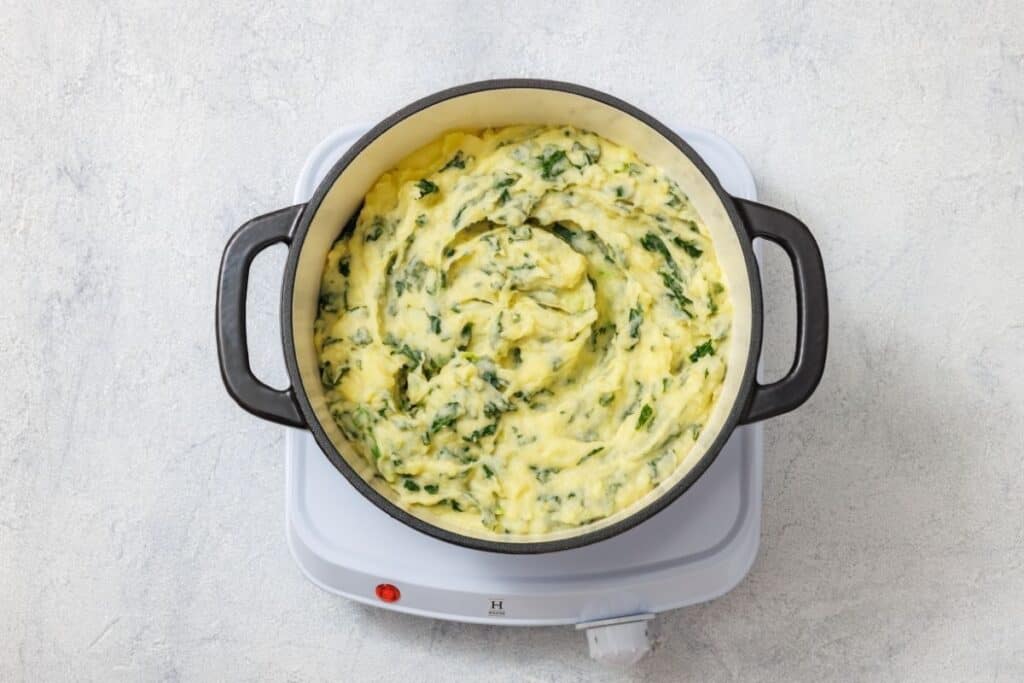 Creamy mashed potatoes with green herbs in a black pot on a white electric hob.