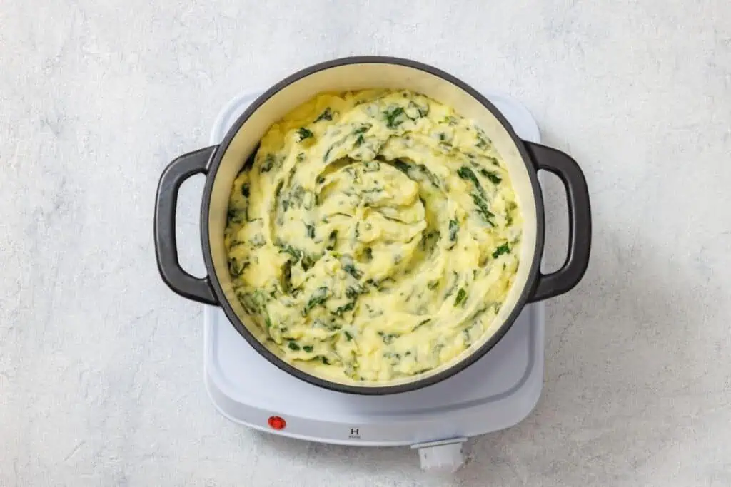 Creamy mashed potatoes with green herbs in a black pot on a white electric hob.
