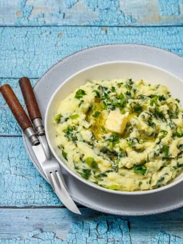 A bowl of Colcannon with Kale with parsley and melting butter on top, placed on a gray plate with a fork and knife on a rustic blue wooden table.