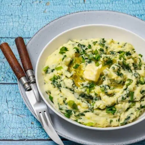 A bowl of Colcannon with Kale with parsley and melting butter on top, placed on a gray plate with a fork and knife on a rustic blue wooden table.