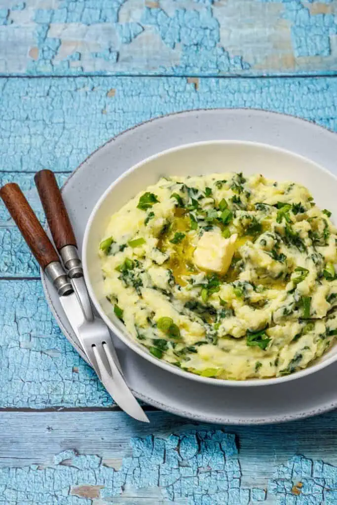 A bowl of Colcannon with Kale with parsley and melting butter on top, placed on a gray plate with a fork and knife on a rustic blue wooden table.