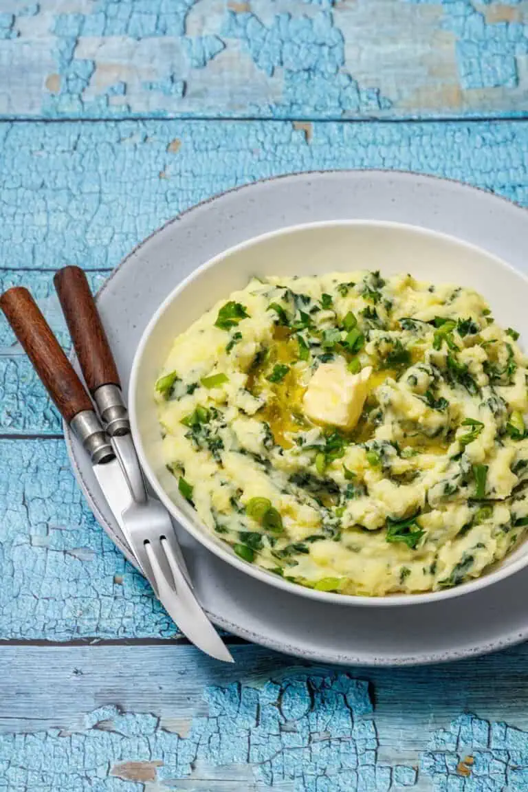 A bowl of Colcannon with Kale with parsley and melting butter on top, placed on a gray plate with a fork and knife on a rustic blue wooden table.