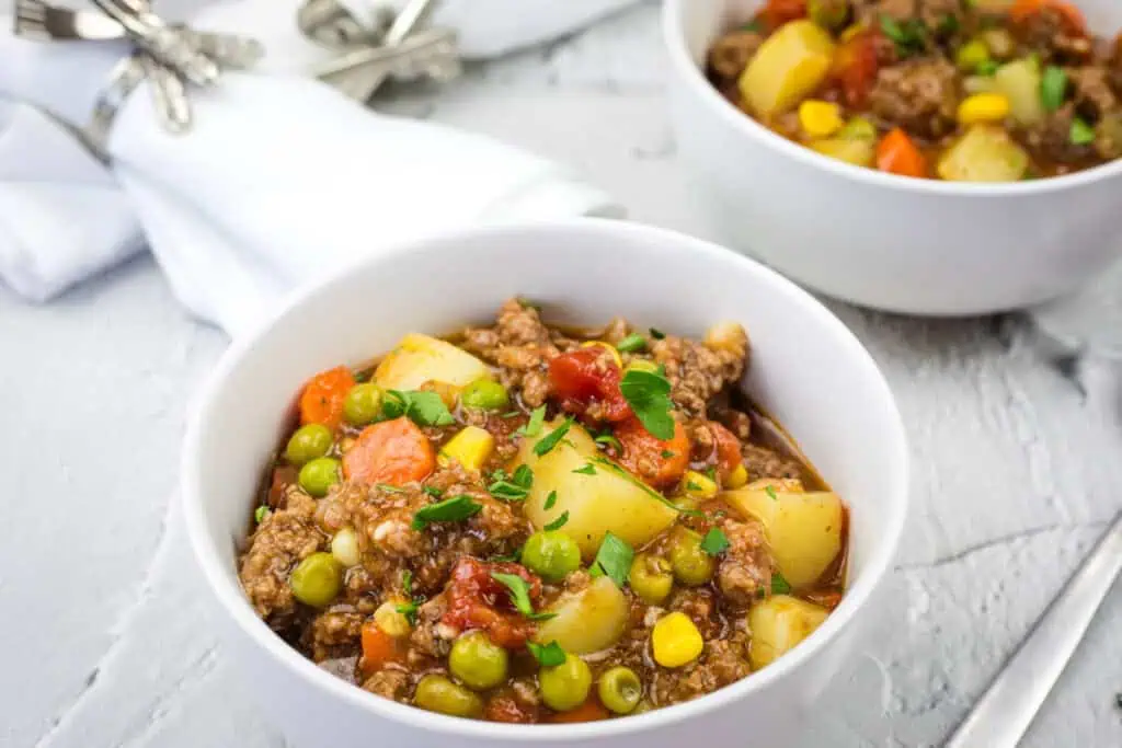 Two bowls of vintage hamburger stew with chunks of potato, carrots, peas, corn, and parsley garnish.