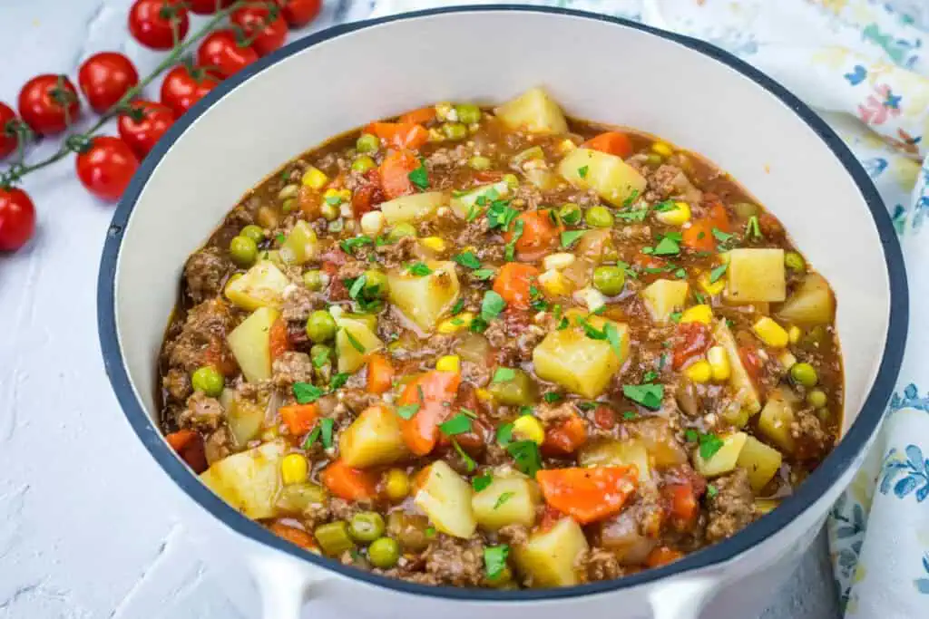 A pot filled with a vintage hamburger stew, featuring chunks of potatoes, carrots, peas, and corn in a rich broth. Cherry tomatoes are visible in the background.