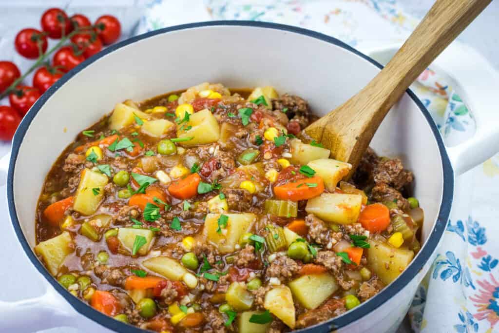 A pot of Vintage Hamburger Stew with diced potatoes, carrots, peas, corn, and tomatoes, garnished with chopped herbs, and a wooden spoon.