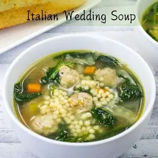 A bowl of Italian wedding soup with meatballs, pasta, spinach, and carrots. A slice of bread is visible in the background.